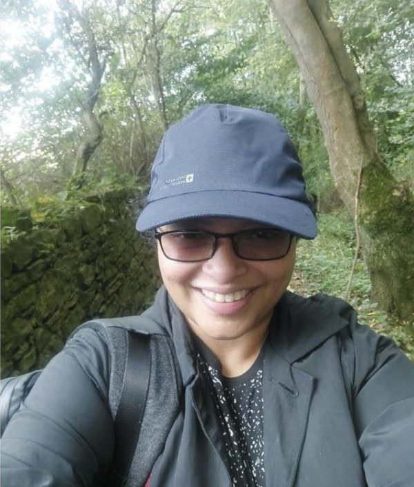 A selfie of a woman wearing a black cap and black sunglasses smiling and standing in front of trees